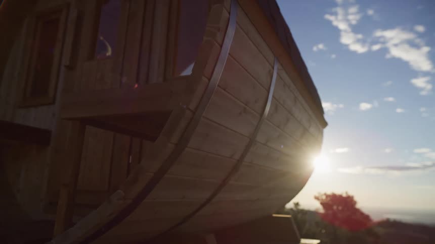 Sun flares over the side of a unique wooden barrel cabin set against a beautiful blue sky