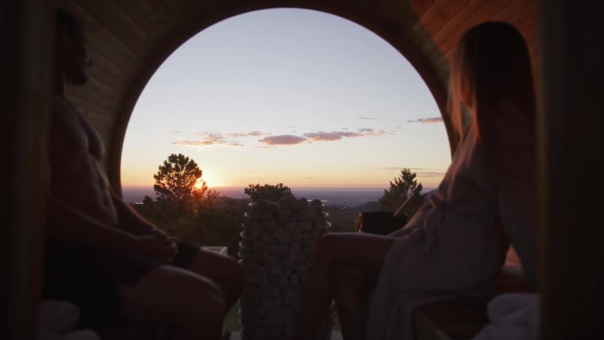 A couple relaxes inside a rustic barrel sauna, enjoying the panoramic view of the mountains as the sun sets