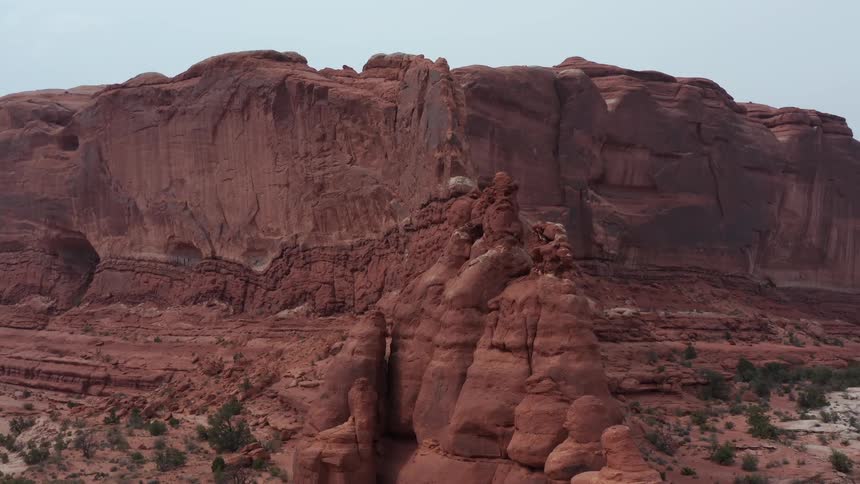 A slow aerial pullback reveals the grand scale of a towering red rock butte and sandstone cliffs in a vast, arid desert landscape