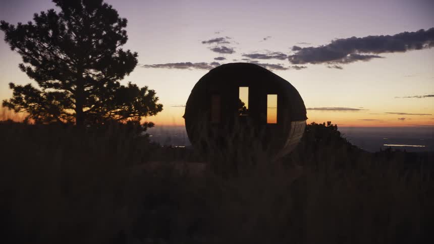 A unique pod cabin sits silhouetted on a grassy mountainside, with the warm colors of sunset reflecting in its windows