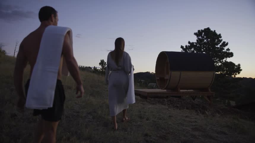 A man and woman walk up a grassy hill towards a rustic barrel sauna at twilight in slow motion