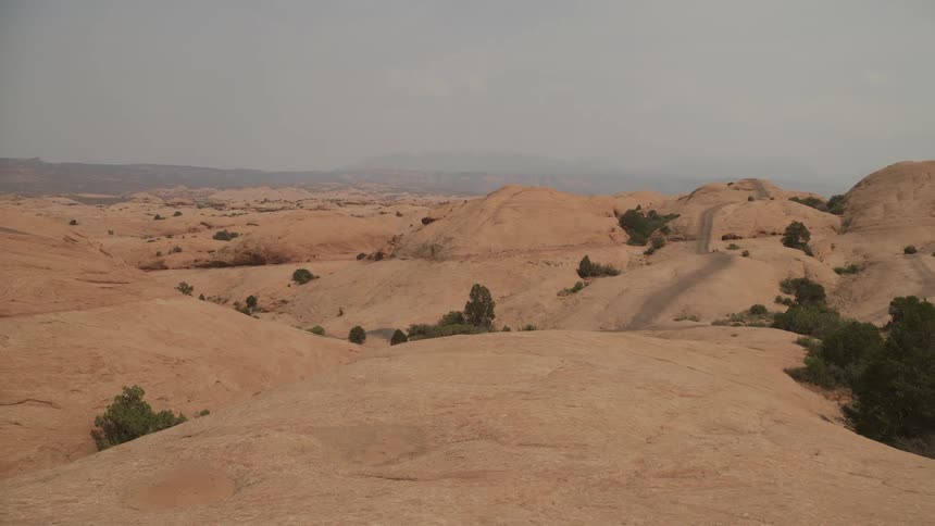 A wide, static view of a vast and arid slickrock desert landscape with a winding trail under a hazy sky