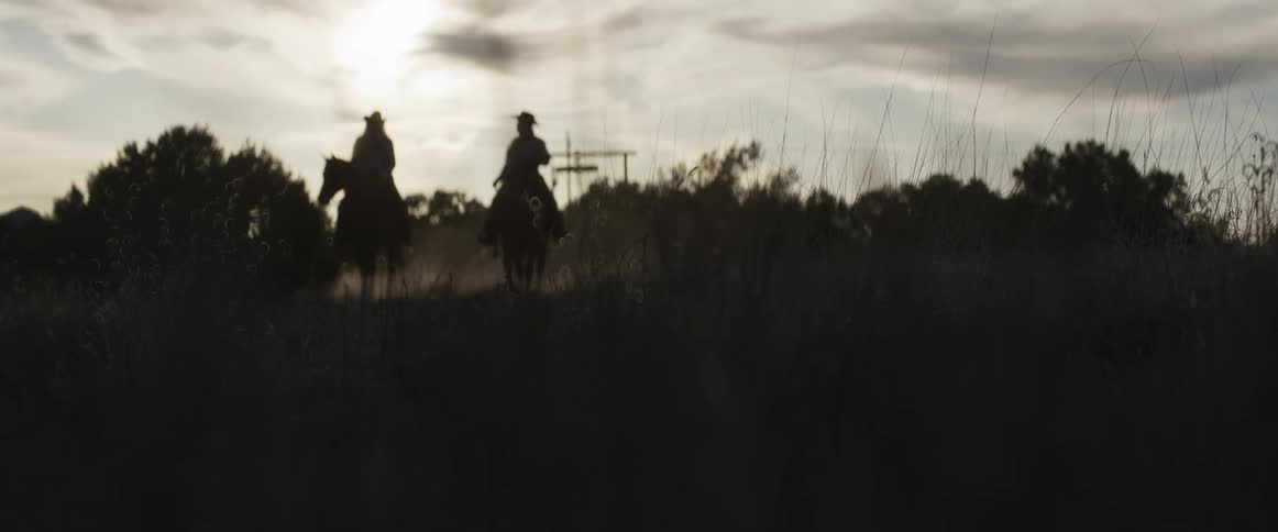 A moody, silhouetted low-angle shot of two cowboys on horseback in a dusty field at sunset