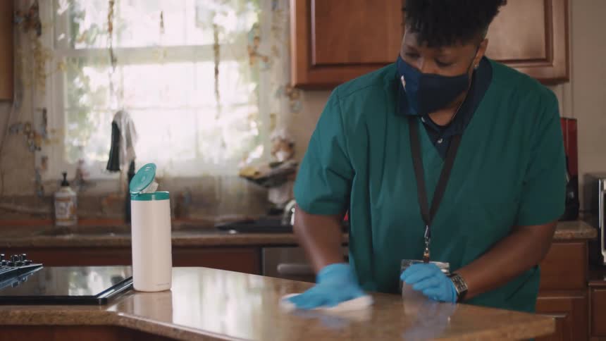A home health aide wearing a face mask and gloves carefully sanitizes a kitchen countertop with a disinfecting wipe in slow motion