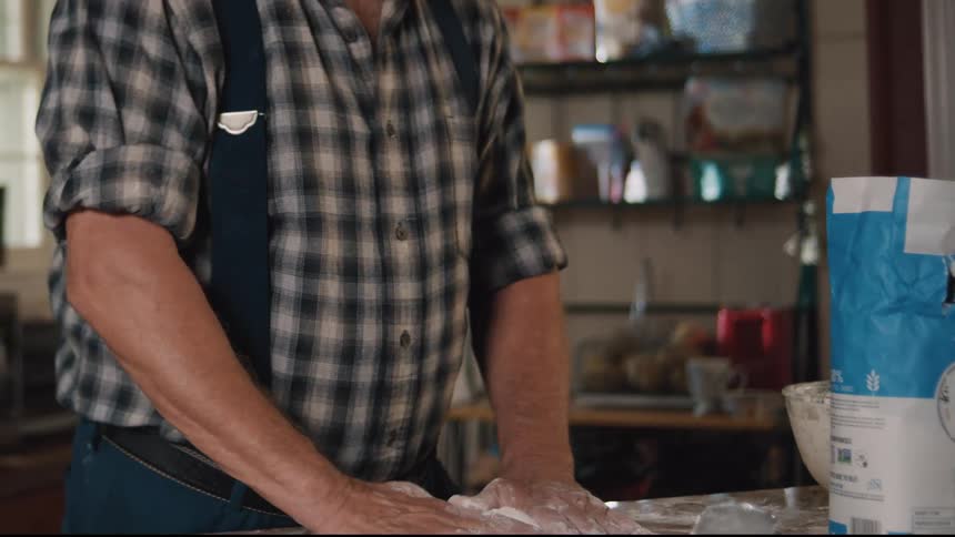 An older man in a plaid shirt and suspenders carefully kneads bread dough in a rustic country kitchen