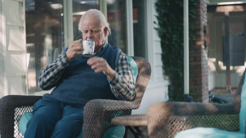 A pensive senior man in a sweater vest relaxes in a wicker chair on the porch, thoughtfully drinking from a mug