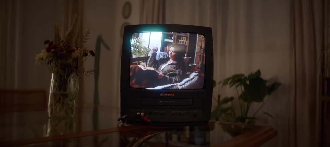 A slow dolly shot pushes in on an old CRT television playing a VHS home movie of a young woman relaxing and reading a book