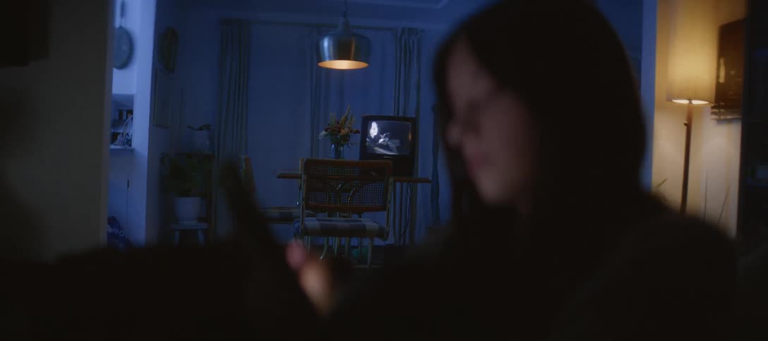 A woman in the dark is illuminated by her smartphone while a classic movie plays on a retro TV in the background