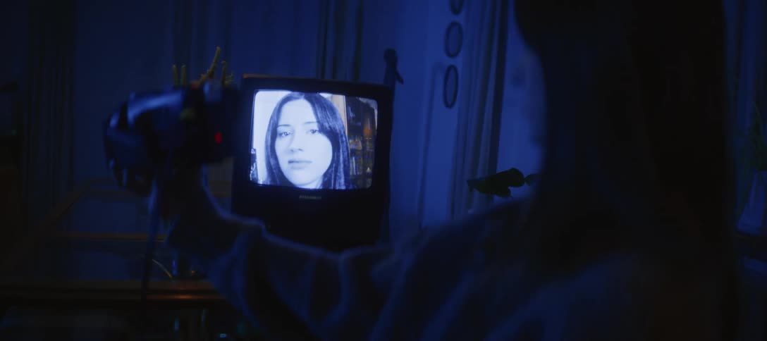 An over-the-shoulder shot of a person filming their black and white self-portrait on a retro television screen with a camcorder in a dark room