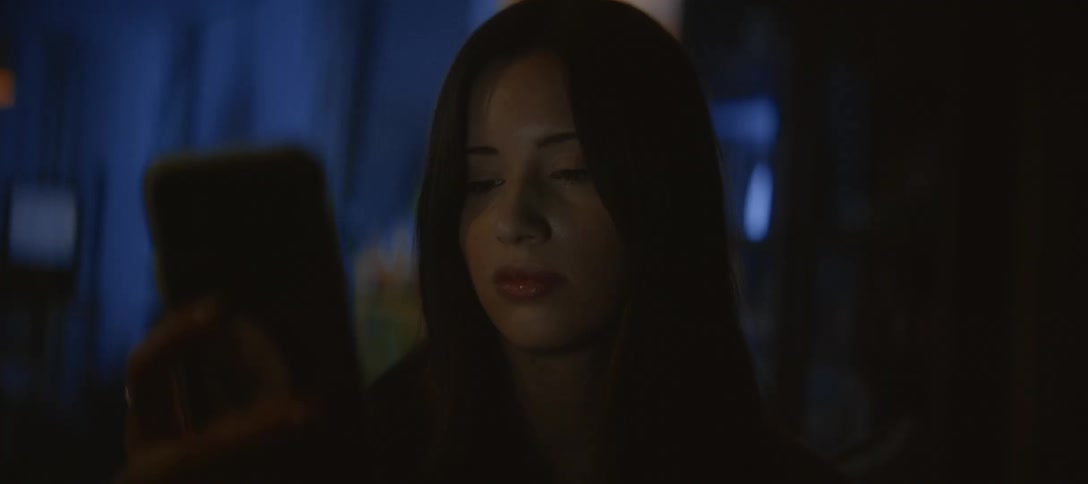 A close up on a young woman's face as she looks down at her phone, illuminated by the screen's glow in a dark room