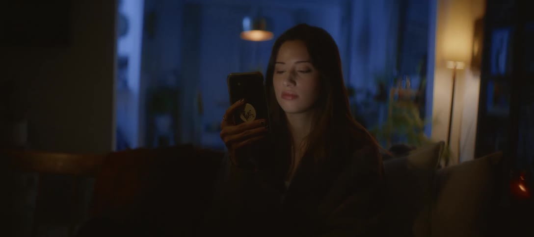 A young woman relaxes on her cozy couch in a dark room, her face illuminated by the glow of her smartphone as she scrolls