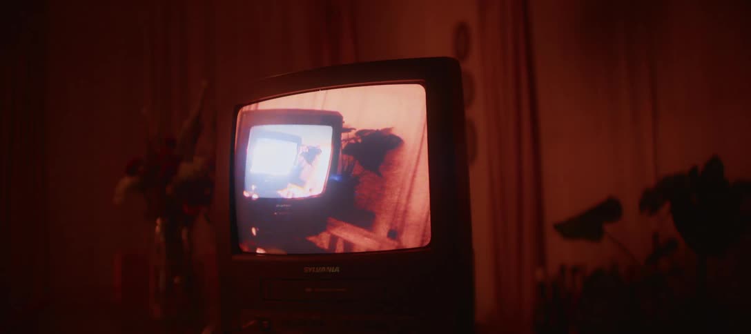 A retro television screen flickers with an old home video, illuminating a dark room with shifting colored lights
