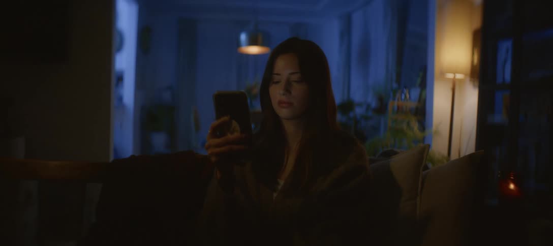 A young woman scrolls on her smartphone in a dark living room at night, her face lit by the screen's glow