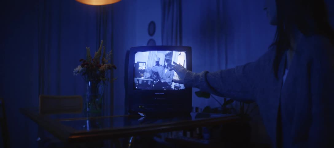 In a dark, blue-lit room, a woman's hand slowly reaches out to touch an old black and white home video playing on a vintage television