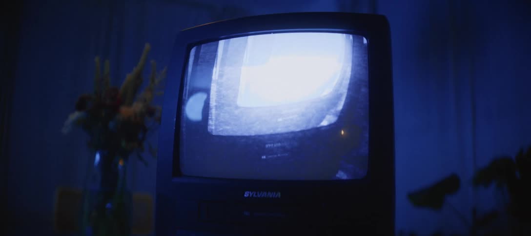 A retro tube television plays a distorted cartoon, casting a blue glow in a dark, moody room