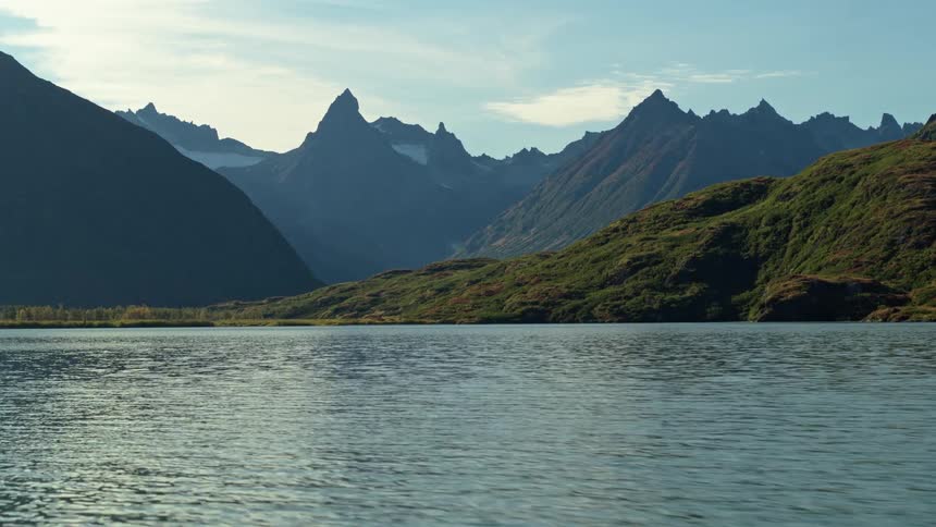 Static shot of a tranquil mountain lake with gentle ripples on the water, surrounded by rolling green hills and jagged, snow-capped peaks in the background