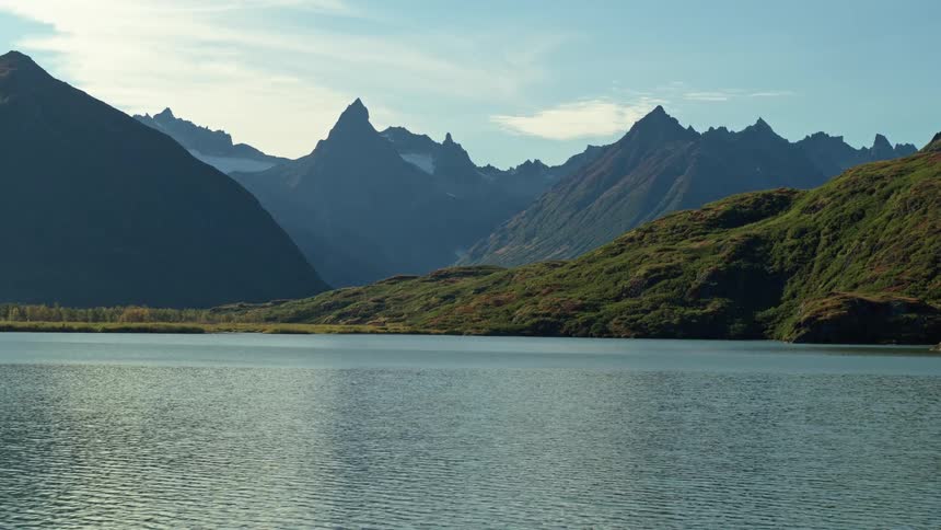 A scenic view of a tranquil lake with rolling green hills leading up to a dramatic and jagged mountain range in Alaska