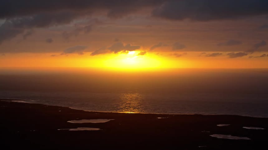 A brilliant golden sun sets over a calm ocean, casting a shimmering reflection on the water and silhouetting the coastline