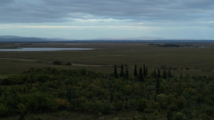 Aerial view slowly rises over a vast green tundra landscape with distant lakes and mountains under a cloudy sky