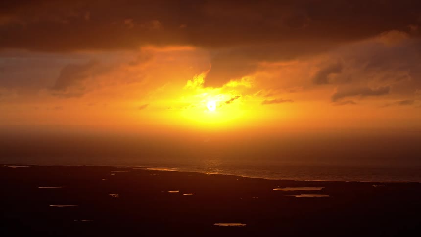 A dramatic golden sunset burns brightly through the clouds over the ocean and a silhouetted coastline