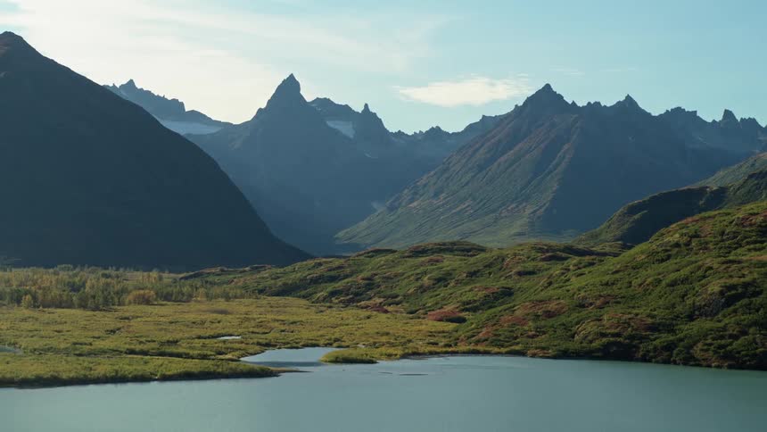 An epic aerial shot pulls back to reveal a majestic mountain range towering over a tranquil lake and lush valley