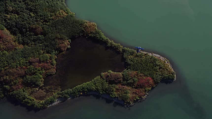 Static top-down aerial view of a seaplane docked on a remote, tranquil peninsula with lush autumn foliage and a small pond