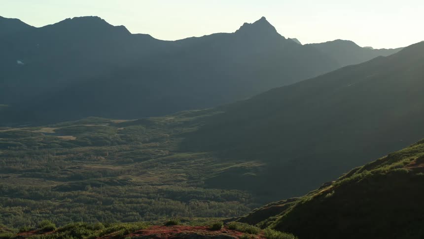 Epic pull-back shot reveals a vast mountain valley with rolling green hills and silhouetted peaks at sunrise