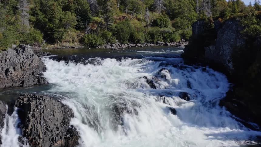 An aerial view slowly advances over a river toward a powerful waterfall cascading over rocks in a lush, green forest