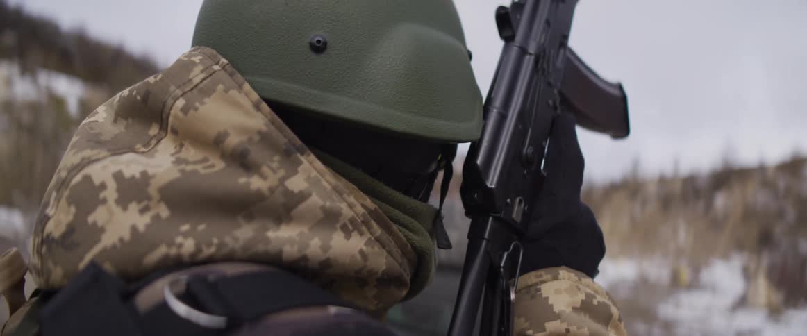 A close-up over-the-shoulder shot of a soldier in winter camouflage holding a rifle in a snowy environment