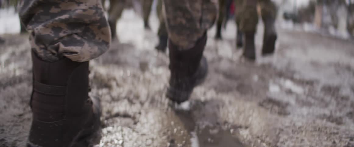 A gritty low-angle slow-motion shot follows soldiers in combat boots as they march through muddy, slushy terrain