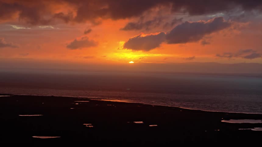 A brilliant orange sunset illuminates a cloudy sky over the ocean and a dark, silhouetted coastline with small reflective pools