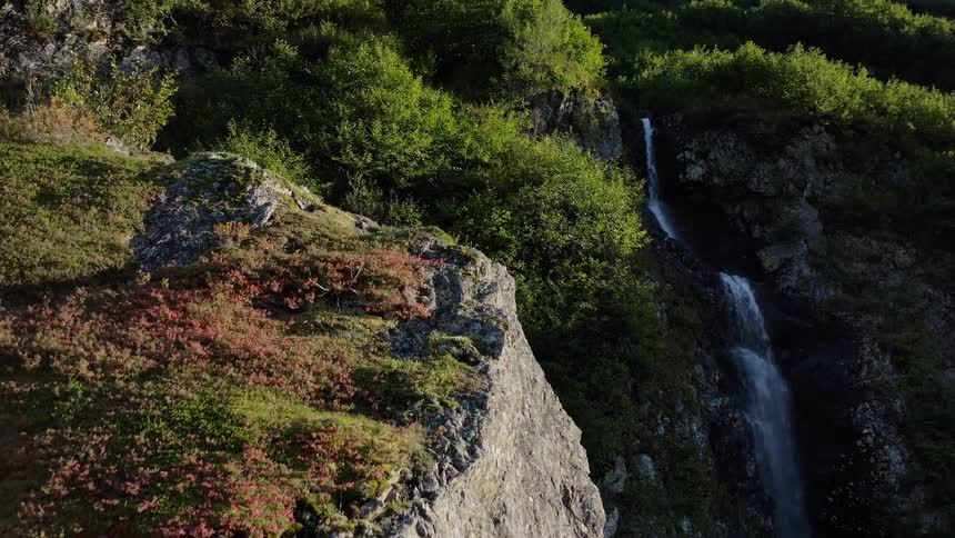 A slow aerial ascent reveals a picturesque waterfall flowing down a rocky, tree-covered mountain slope