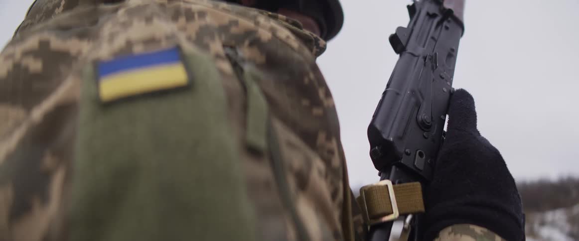 A close-up shot of a Ukrainian soldier in camouflage holding a rifle, with the flag of Ukraine on their shoulder