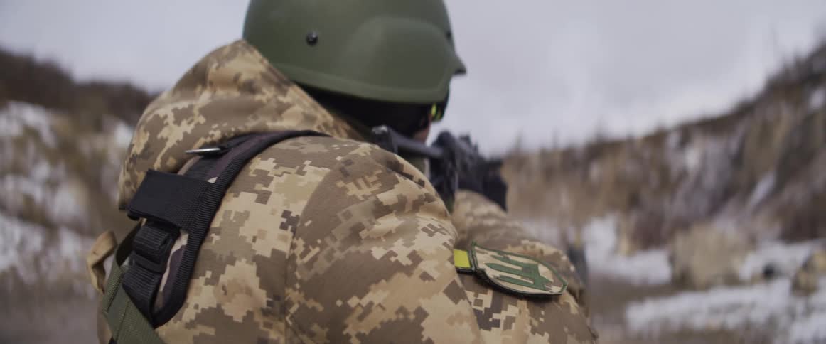 Over-the-shoulder slow-motion shot of a Ukrainian soldier in a helmet and camouflage uniform firing a rifle at a snowy outdoor range
