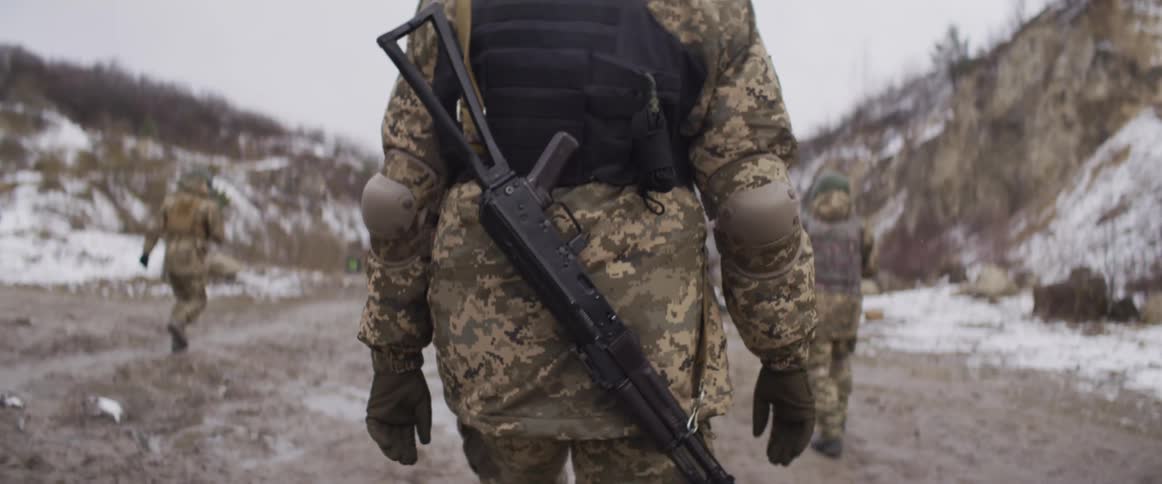 Slow motion shot from behind of a Ukrainian soldier in camouflage with a rifle walking in a snowy quarry with his unit
