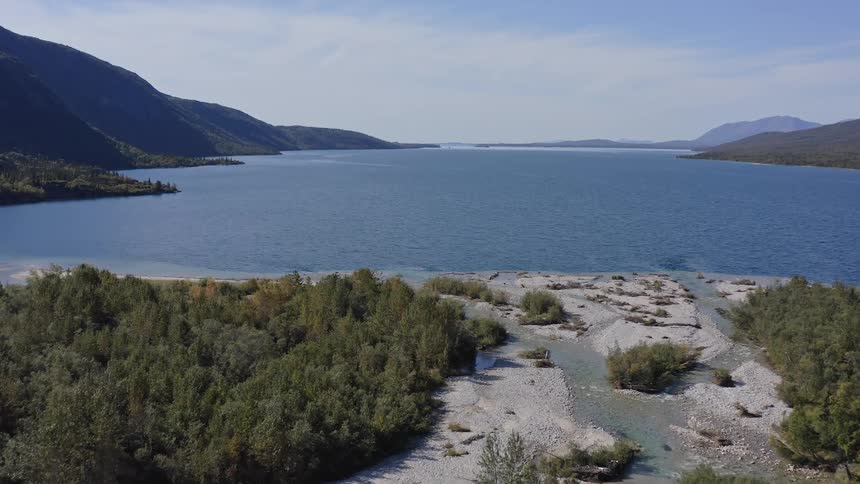 A slow and majestic aerial flight over a rocky river delta and green forest towards a vast, serene mountain lake on a sunny day