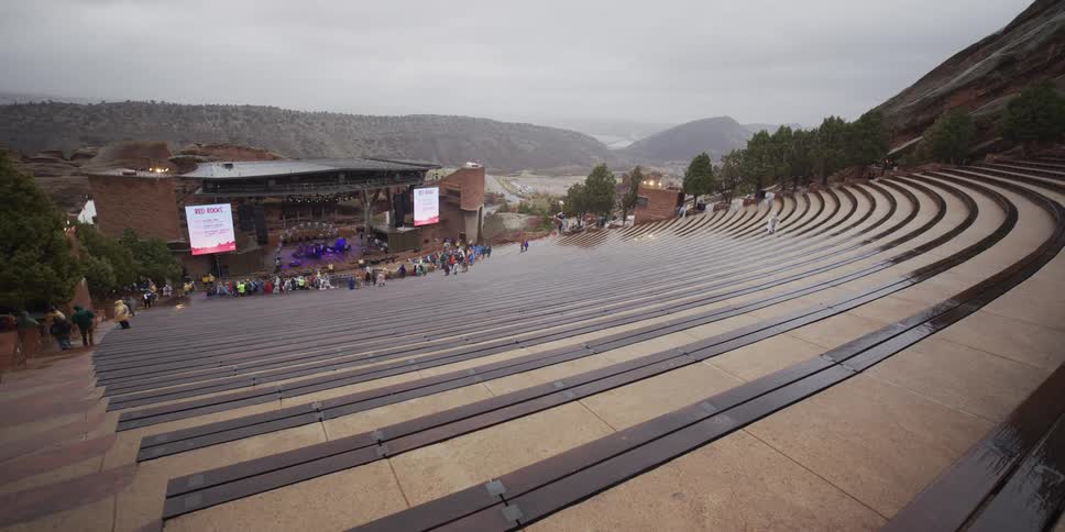 A wide static shot captures the empty, rain-slicked seats of Red Rocks Amphitheatre on a cloudy day as a band prepares on stage