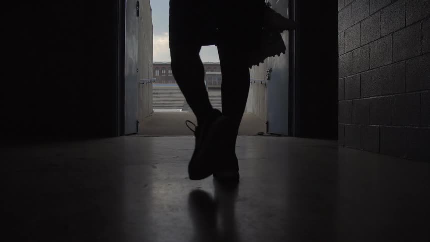 Man holding a football and cleats walks down a long corridor
