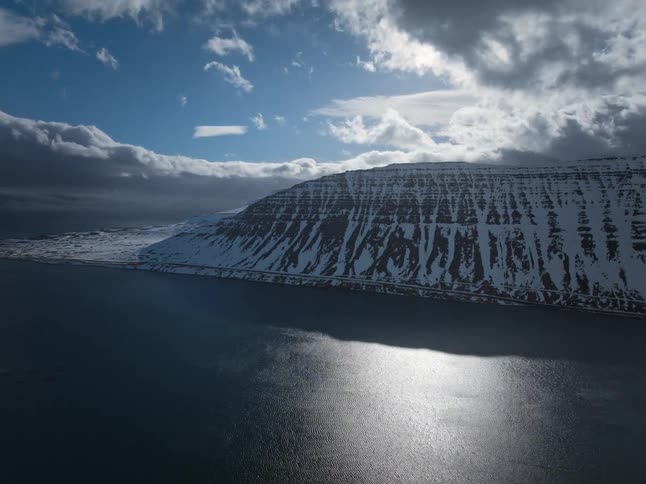 A stunning aerial timelapse captures sunlight and clouds moving across a vast fjord and a dramatic, snow-covered mountain range