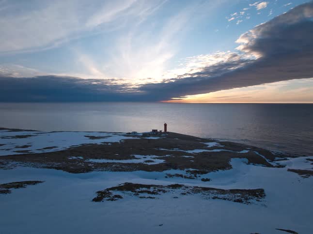 Slow aerial pullback reveals a remote red lighthouse on a snowy coastline during a beautiful sunset