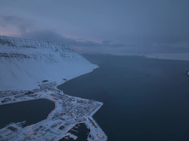 Aerial view of a snowy Icelandic coastal town with glowing lights nestled in a fjord at twilight