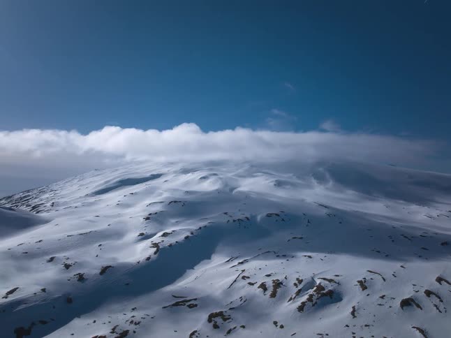 A majestic aerial timelapse shows clouds sweeping across a vast, snow-covered mountain under a bright blue sky