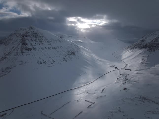 An aerial view of a road winding through a snow-covered mountain valley as sun rays break through dark clouds
