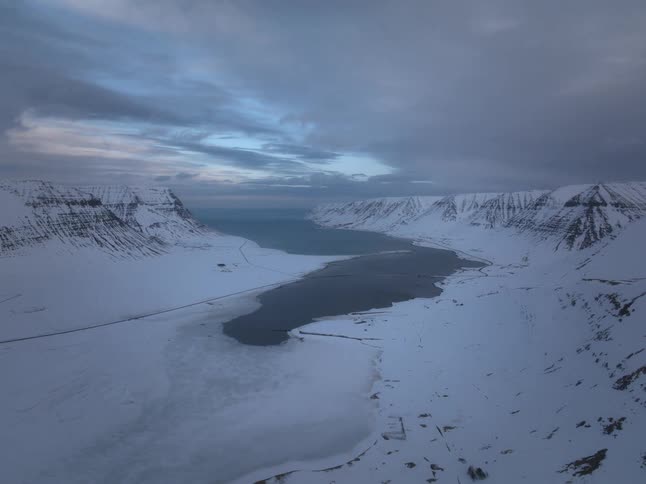 A slow aerial pullback reveals a vast and desolate snow-covered fjord leading to the ocean under a dramatic twilight sky