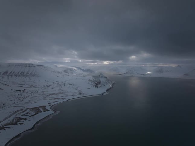 A dramatic aerial view shows sunbeams breaking through moody clouds over a vast, snow-covered fjord and mountains