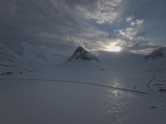 A majestic time-lapse captures the sun breaking through dramatic clouds to illuminate a vast, frozen mountain landscape