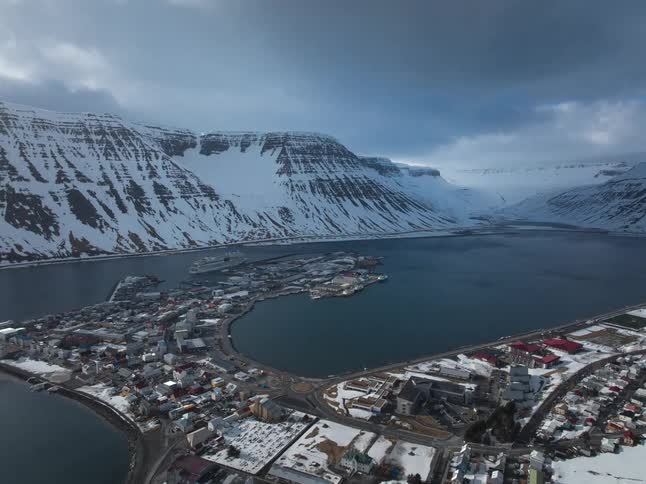 A dramatic aerial hyper-lapse flies over the snowy town of Ísafjörður, Iceland, nestled within a massive fjord as clouds race across the sky