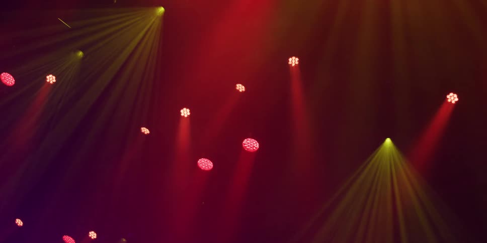 Stage spotlights in red and yellow shine down through a hazy, dark atmosphere