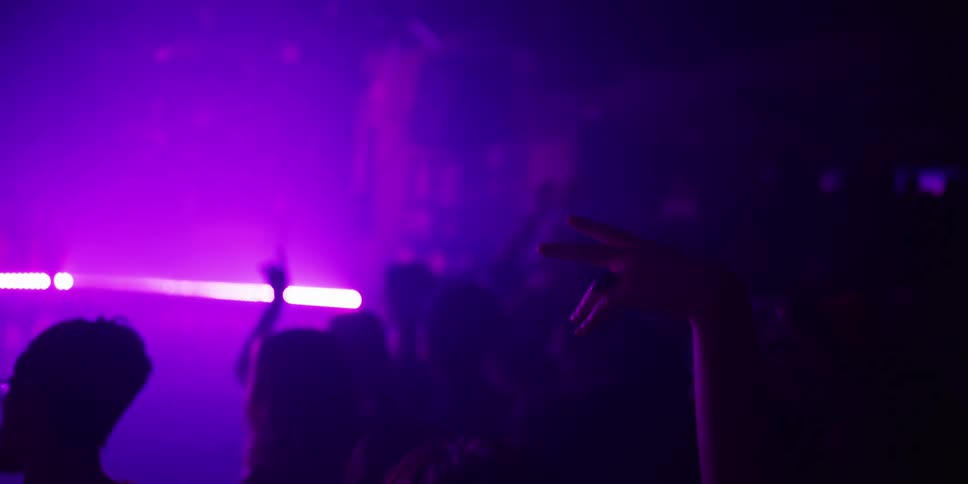 A silhouetted hand sways to the music in slow motion as a vibrant crowd dances under purple lights at a nightclub