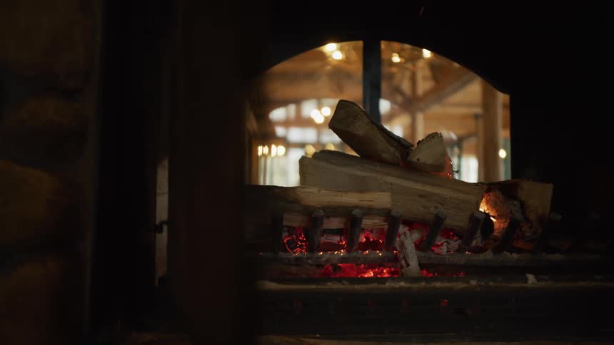 Logs smolder over glowing red embers in a cozy stone fireplace, reflecting the warm interior of a rustic lodge